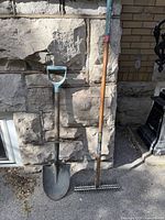 Full view of spade shovel and dethatching rake leaning against stone wall