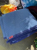 Folded blue quilted moving blanket with red tag and visible white paint splatters
