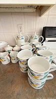 Group view of stacked mugs, two lidded pots, sugar bowl and creamer on countertop