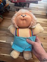 Front view of Koosas plush cat doll showing face, mane, rainbow suspenders, blue shorts, collar tag