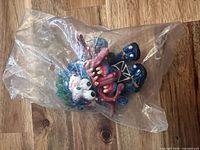 Figure sealed in clear plastic bag, front view showing blue limbs, pink tentacles, white face