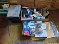 Overall lot showing plastic storage bin, cardboard box and stacked office supplies including label maker, labels, envelopes