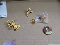 All four items arranged on surface: two angel pins, flag pin in sleeve, portrait button