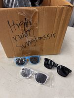 Open carton with numerous bagged sunglasses plus three sample pairs (blue/yellow, white, black) on table