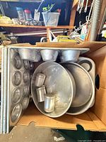 Box containing assorted aluminum bakeware showing muffin pans, cake pans, popover molds and dredgers