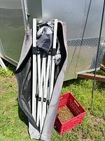 Folded white metal scissor-frame canopy inside gray carry bag next to red milk crate