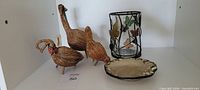 Three wicker duck figurines, glass vase with leaf metal frame, ceramic plate displayed on shelf