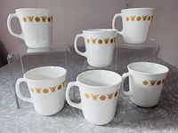 Six white mugs with Butterfly Gold pattern displayed on acrylic risers
