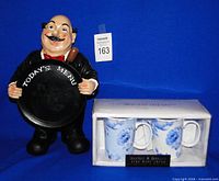 Front view of resin maître d' figure with Today’s Menu board beside boxed pair of bone china mugs