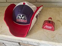 Front view of navy and white mesh Brock Badgers hat beside red Brock University cowbell