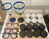 Group shot showing 2 blue-rimmed goblets, 3 pairs reading glasses, stacked white and black beer can carriers