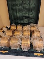 Open green velvet case showing brass napkin rings inside, some wrapped in plastic