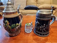 Three lidded beer steins—large Gertz knight scene, miniature BMF glass, medium Thewalt hunter scene—group view