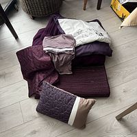 Stacked bedding items on floor: quilt, fuzzy blanket, folded shams, accent pillow, table runner visible