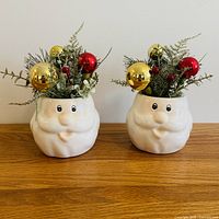 Front view of two ceramic Santa head decorations with greenery and ornaments