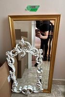 Rectangular gold-frame mirror standing upright behind ornate white-framed mirror