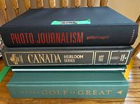 Stack of four hardcover books showing titles Photo Journalism, Canada Heirloom Series, Where Golf Is Great with additional yellow book partially visible