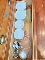 Drawer view showing four rectangular canisters with lids, cylindrical canister, pink strainer, ceramic cups, glass clamp jar