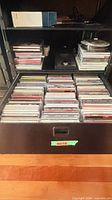 Drawer containing rows of audio CDs in jewel cases; shelves with additional CD stacks