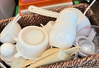 Basket containing assorted white plastic measuring cups, spoons, strainer, beige kitchen tool and clear liquid measuring cup