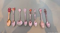 Eight souvenir spoons laid out side by side showing overall styles and crests