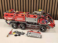 Left side view of partially assembled 6-wheel Lego Technic airport rescue truck with loose parts in foreground