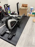 Side view of Fitness Reality rower folded on mat, shows flywheel housing and foot pedals
