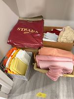 Three containers of assorted folded linens including Strada Italy packaged tablecloth, bright placemats, pink napkins and crochet items