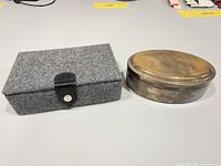 Closed gray felt jewelry box beside closed oval silver-plated box