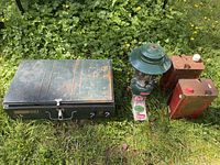 Closed green Coleman 2-burner stove, lantern, two fuel cans, mantle package on grass