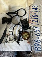 group shot of entire lot showing axe, magnifiers, vise, ring light, loupe, book, yo-yo, licence plates