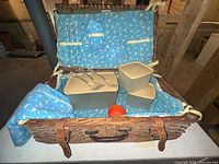 Open wicker picnic basket showing interior lining, Tupperware containers, orange snack cup, utensils