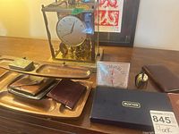 Front view of mantel clock, Taylor instrument, valet tray with wallets, brass bell and Buxton box