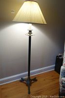 Full view of vintage brass finished metal floor lamp illuminated with pleated fleeced fabric shade, standing against a wall on a hardwood floor.