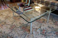 Front-left angle view of the rectangular coffee table with a clear, shaped glass top and white painted iron base set on a floral area rug.