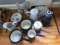 Vintage kitchenware items including enameled pots, ceramic pitchers, a hand-painted bowl, and ceramic candlesticks arranged on wood floor.