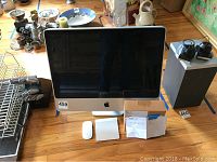 Front view of Apple iMac on wooden floor with Magic Mouse, Magic Trackpad, brown box and invoice sheet visible