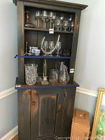 Distressed rustic cupboard showing open shelving with a variety of glassware on the shelves and a single wooden door cabinet below.