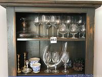 Shelves displaying glass decanter, various wine glasses, two metal candlesticks, blue and white ceramic bowls, and colorful figurines.