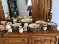 Full set of Mikasa Grande Ivory dinnerware displayed on wooden sideboard showing plates stacked with teacups, creamers, sugar bowl and bowls visible, including some floral patterned pieces at the edge.