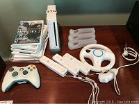 Full display of Nintendo Wii console, Wii remotes, Wii wheel controller, Wii nunchuck controller, Xbox 360 controller, and stack of Wii games.