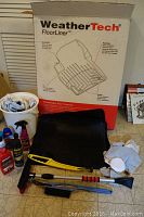Photo of the original WeatherTech FloorLiner box, black molded floor mats, car wax and cleaning products, yellow and black handled cleaning tools and microfiber cloths on floor.