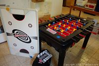 Multi-game table frame with foosball top installed, showing plastic handles and player rods with blue, yellow, and red players. Air hockey top and storage bag beside it.