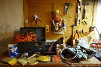 Setup showing Plano toolbox filled with an orange extension cord, cordless drill, drill bits, white brushes, and various small hand tools on a workbench with pegboard background.