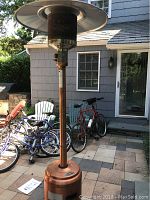Full view of the Fire Sense propane patio heater standing outside beside bikes and house wall, showing overall structure and wheels.