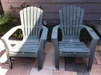 Two green plastic Adirondack chairs positioned side by side on outdoor patio tiles against siding, showing surface dirt and wear.