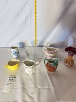 Group shot of all ceramic creamers, clear vase and lot tag with measuring tape for scale