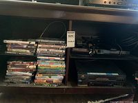 Samsung Blu-ray player and JVC VCR on shelf beside stacks of DVDs