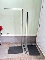 Two disassembled black metal garment racks with bases and poles leaning against wall