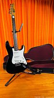 Full view of black Oscar Schmidt electric guitar on stand with open hard shell case